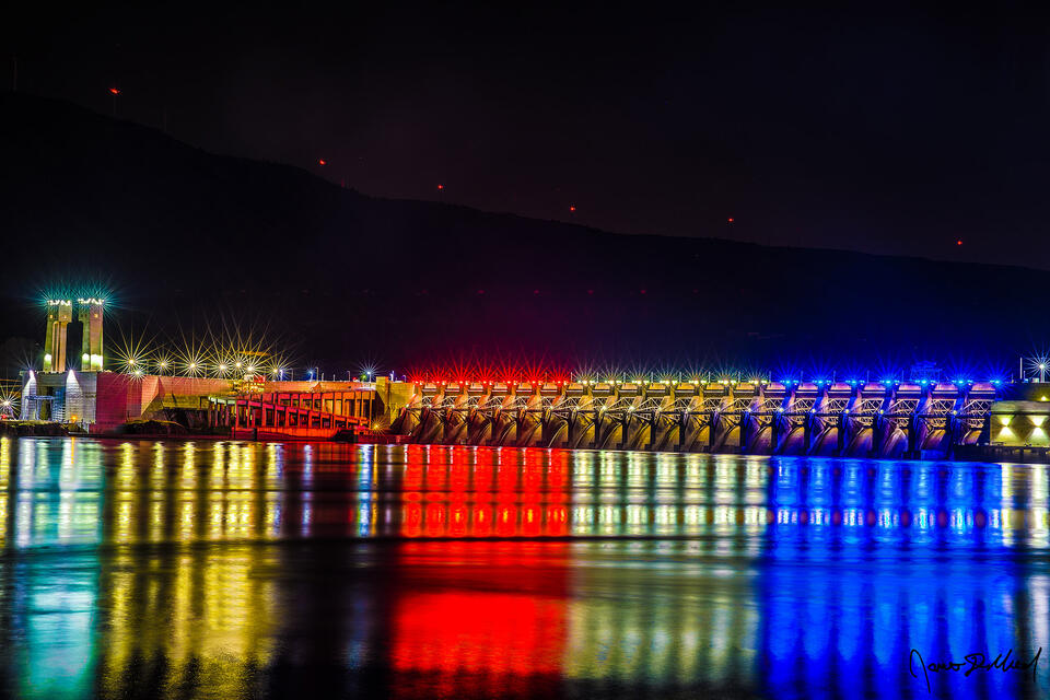 Red, white and blue lights of the John Day Dam reflect off the waters of the Columbia River at night as the red lights of the Windmills line the slopes above.