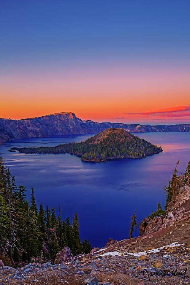 Warm sunset colors light the skies over Crater Lake and Wizard Island from Discovery Point as the alpenglow illuminates Llao Rock and part of the water.