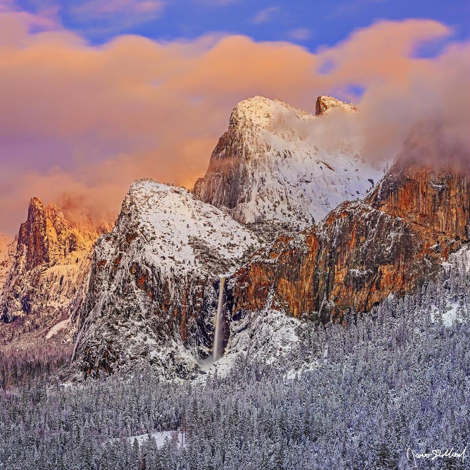 Clouds light up with alpenglow as they cover the top of Cathedral Rocks at Sunset. Bridalveil Fall tumbles over the granite cliffs towards Yosemite Valley.