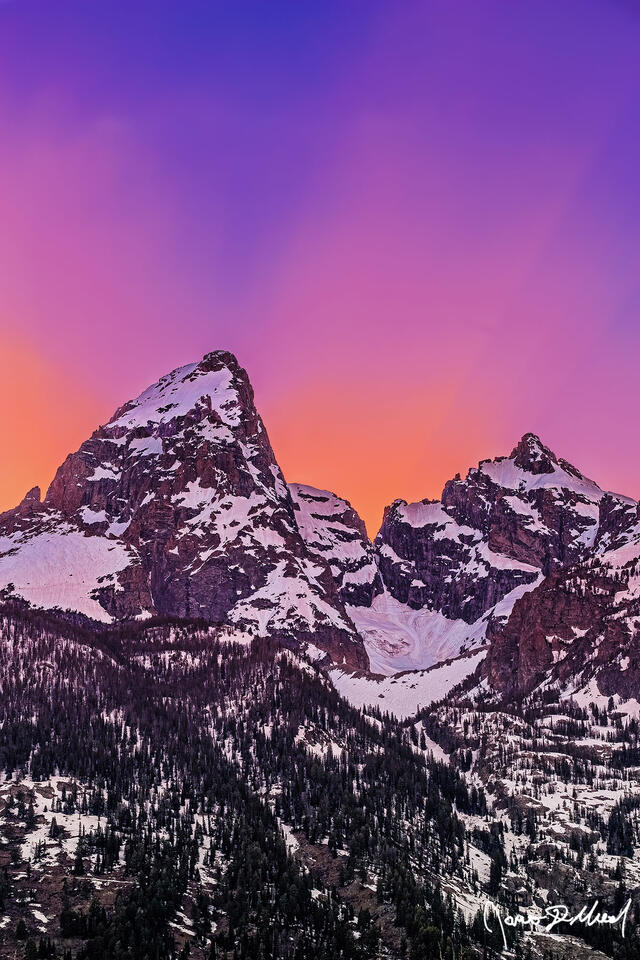 The skies ignite with soft glowing hues at sunset as marvelous beams of pastel light rise up from behind Grand Teton and Mount Owen and into the heavens above.