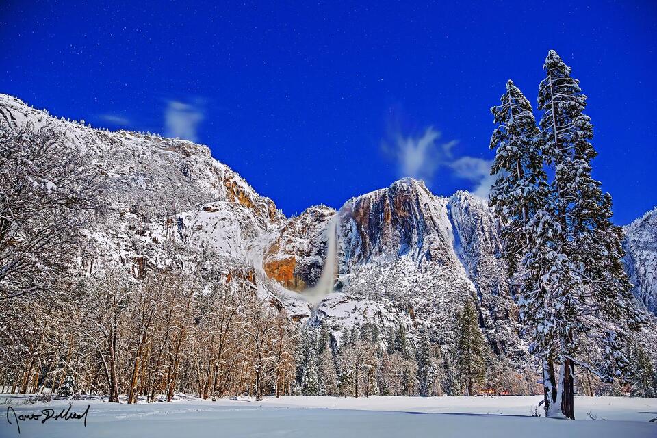 Winter moonlight illuminates the night sky, Yosemite Falls and the granite cliffs above Yosemite Valley as snow blankets Cook's Meadow and the valley below.