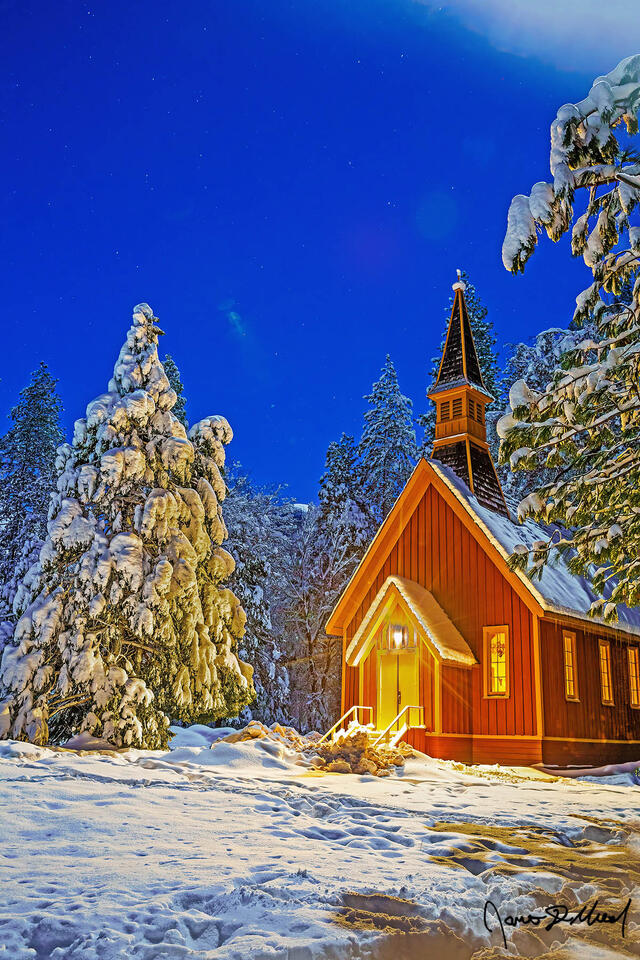 Snow blankets the surrounding meadow, trees and rooftop of Yosemite Valley Chapel on a cold winter night as moonlight illuminates the night sky high above.