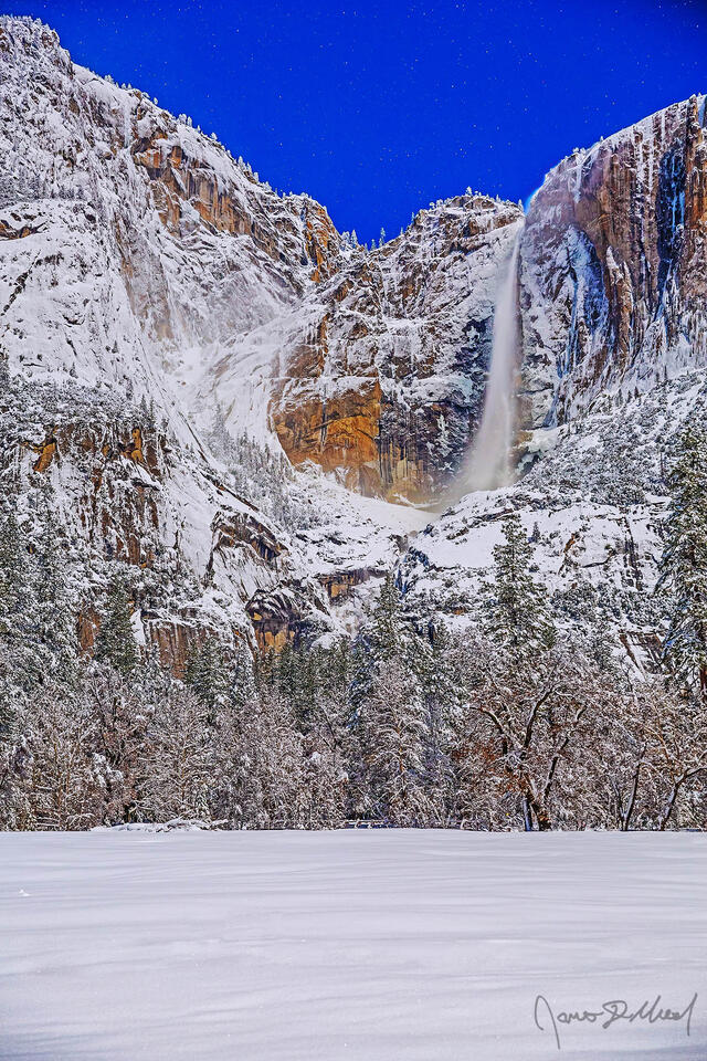 Moonlight illuminates the night sky, Yosemite Falls and the granite cliffs above Yosemite Valley in the winter as snow blankets Cook's Meadow and the valley.