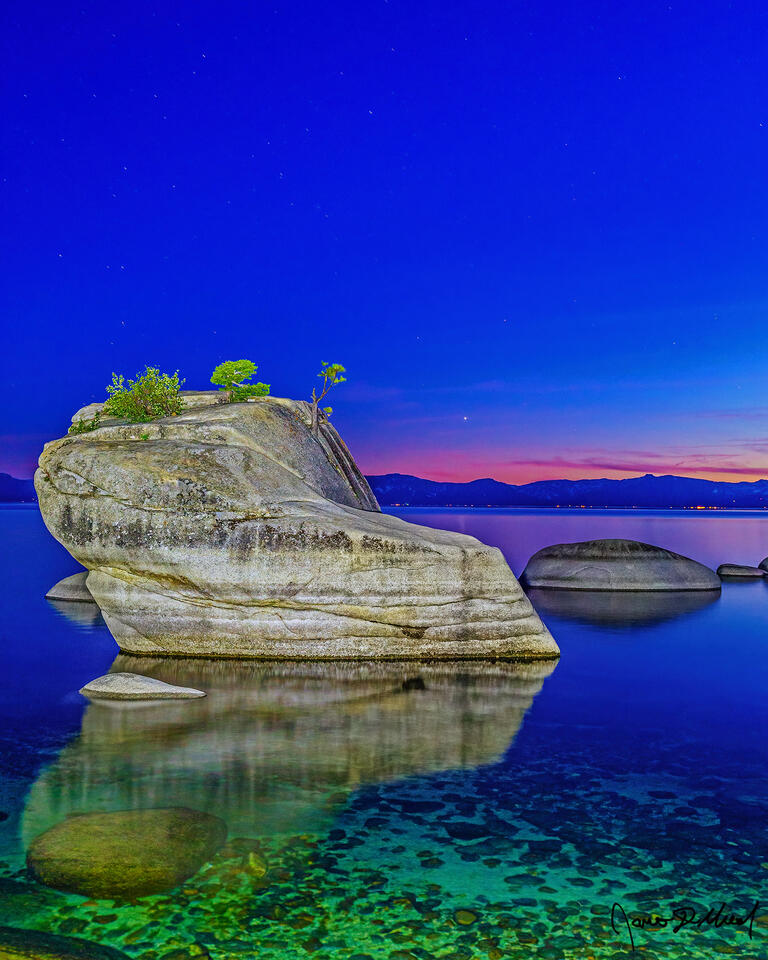 Slight alpenglow hangs on along the horizon, reflecting off the water over of Lake Tahoe at nautical dusk while Bonsai Rock rises out of the clear waters.