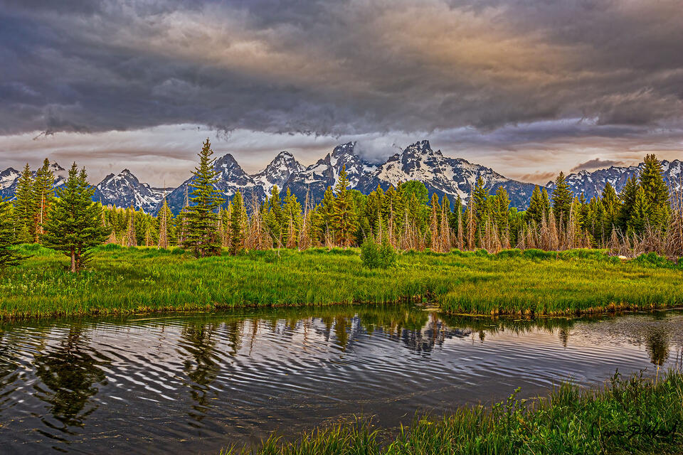 Storm clouds cover the peaks of Grant Teton and Mount Owen as sunrise illuminates the Cathedral Group and the clouds above a branch of the Snake River.