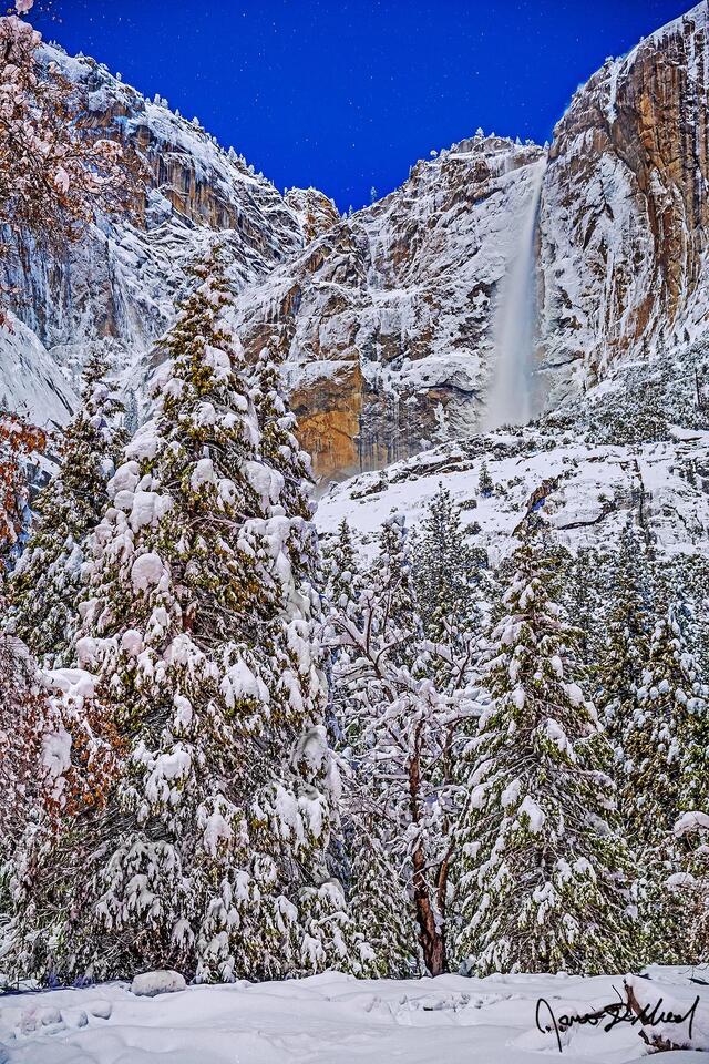 Moonlight illuminates the night sky, Yosemite Falls and the granite cliffs above Yosemite Valley as snow blankets Cook's Meadow and the valley below.