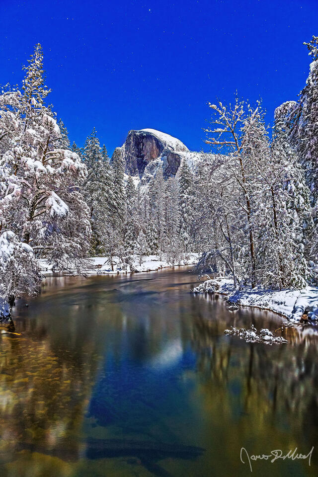 Moonlight illuminates Half Dome as it reflects in the still water of the Merced River. Snow blankets Yosemite Valley and the top of Half Dome as the stars above