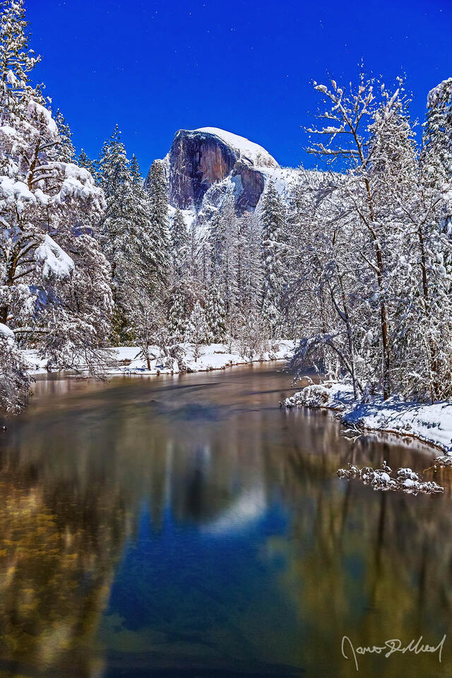Moonlight illuminates Half Dome as it reflects in the water of the Merced River. Snow blankets Yosemite Valley and the top of Half Dome as the stars shine.