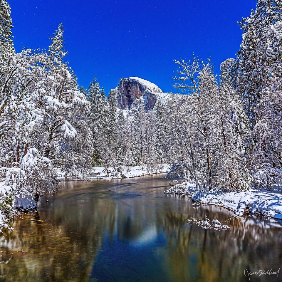 Moonlight illuminates Half Dome as it reflects in the water of the Merced River. Snow blankets Yosemite Valley and the top of Half Dome as the stars shine above