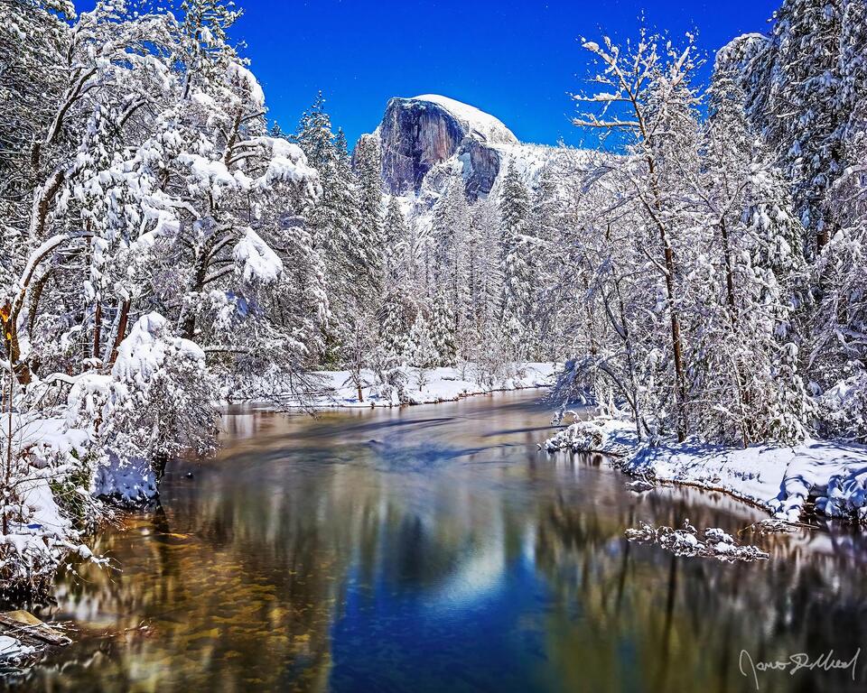 Moonlight illuminates Half Dome as it reflects in the still water of the Merced River. Snow blankets Yosemite Valley and the top of Half Dome as the stars shine