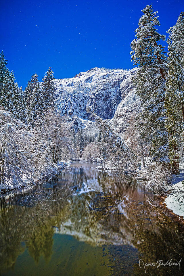 Moonlight illuminates snow covered Yosemite Valley granite cliffs above as the stars shine in the sky; the scene reflects in the waters of the Merced River