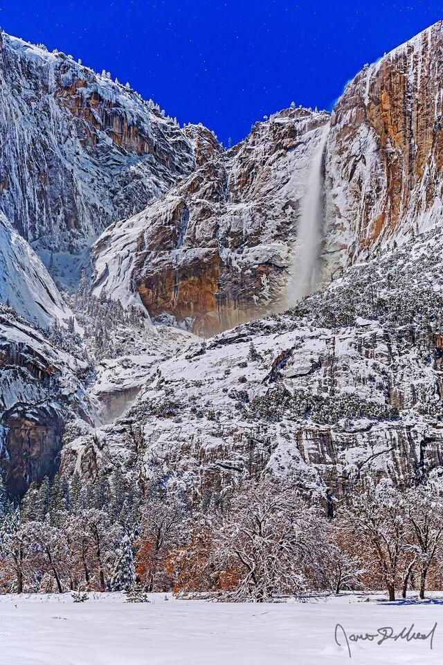 Moonlight illuminates the night sky, Yosemite Falls and the granite cliffs above Yosemite Valley in the winter as snow blankets Cook's Meadow and the valley.