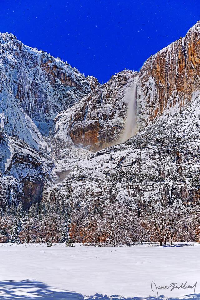 Moonlight illuminates the night sky, Yosemite Falls and the granite cliffs above Yosemite Valley in the winter as snow blankets Cook's Meadow and the valley.