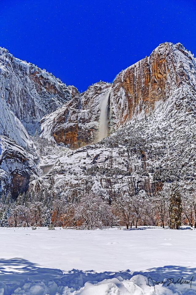 Moonlight lights the night sky, Yosemite Falls and the granite cliffs above Yosemite Valley in the winter as snow blankets Cook's Meadow and the valley below.