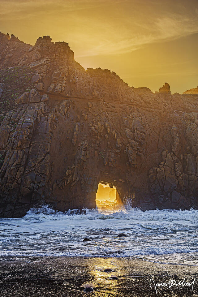 Light beams shine through Keyhole Arch along Pfeiffer Beach in Big Sur as sunset illuminates the skies above.
