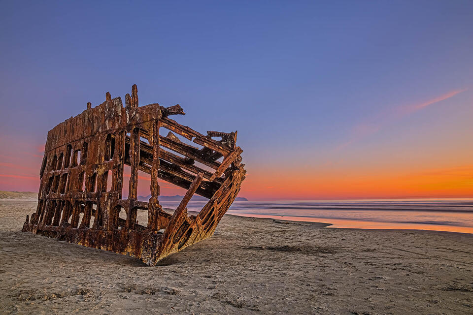 Sunset casts a pastel glow onto skies behind the bow of the Wreck of the Peter Iredale on the Oregon Coast as it lay silent on the beach.