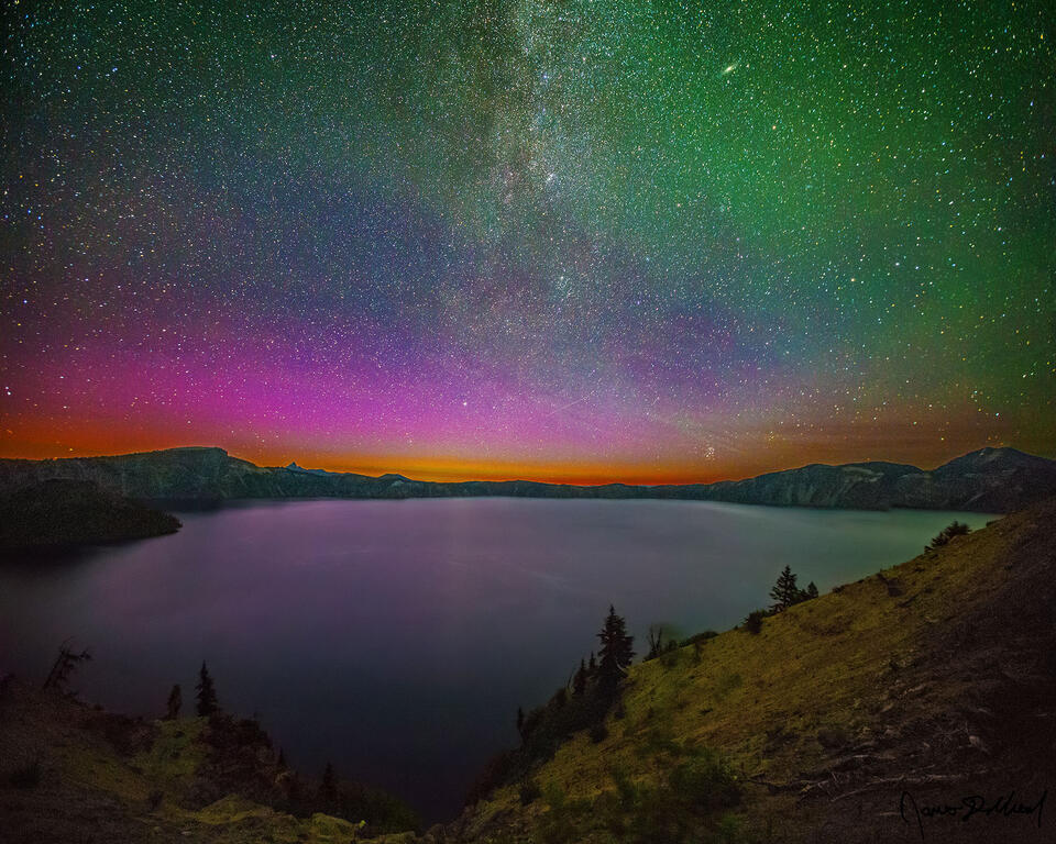 Magentas and greens of the Aurora Borealis light the night sky, faintly reflecting off the waters of Crater Lake as the Milky Way spiral arm rise into the sky.