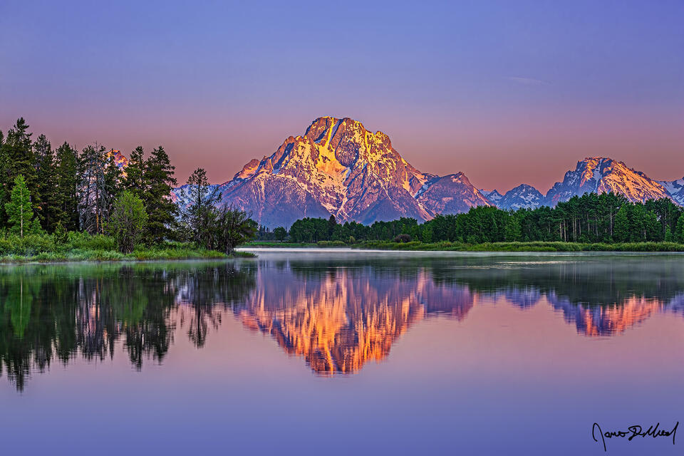 Sunrise illuminates the slopes of Mount Moran, Bivouac Peak and Mount Woodring as the scene reflects in the still waters of the Snake River at Oxbow Bend.