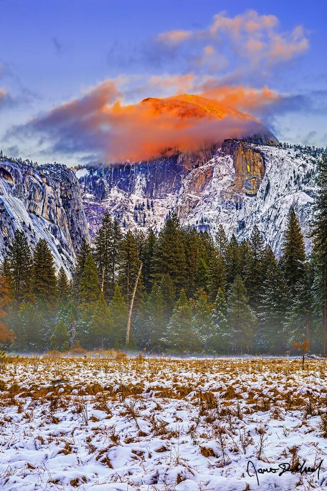 Fiery alpenglow at sunset illuminates Half-Dome at sunset as it rises above the Yosemite Valley floor partially cloaked in clouds.
