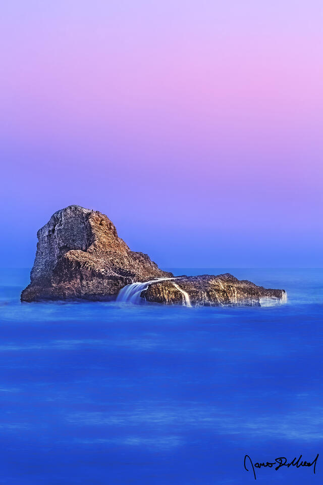 The Pacific Ocean splashes over Pelican Rock at sunset as the Belt of Venus illuminates the skies above as water cascades off the edges of the rock.