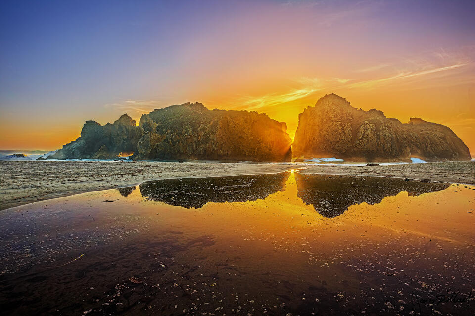 Sea stacks reflect in a pool of water at sunset on Pfeiffer Beach as the Pacific Ocean plunges through an arch opening as sunlight peaks through.