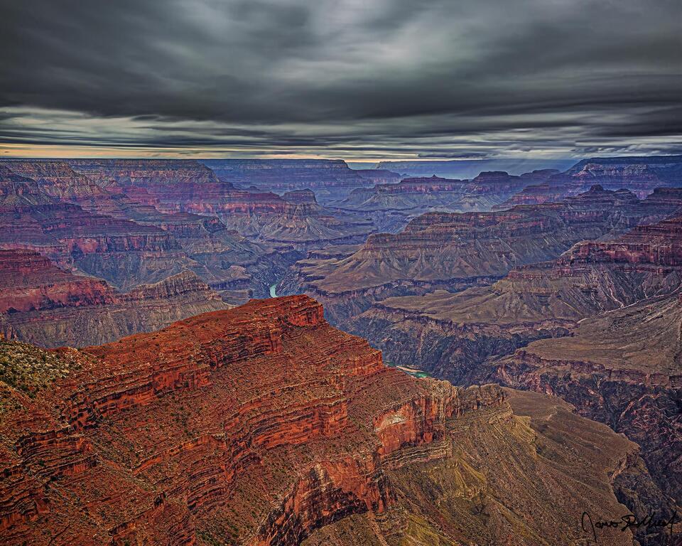 Storm clouds above the Grand Canyon and the Colorado River as the colors of sunset peak through on the horizon as a rainstorm drops onto Powell Plateau.