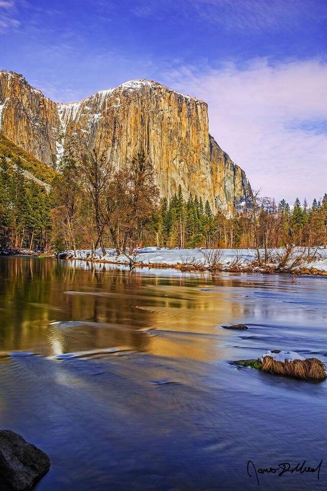 Sunset approaches as colors begin to show in the skies above Yosemite Valley, El Capitan, and the Merced River in the winter while snow blankets the ground.