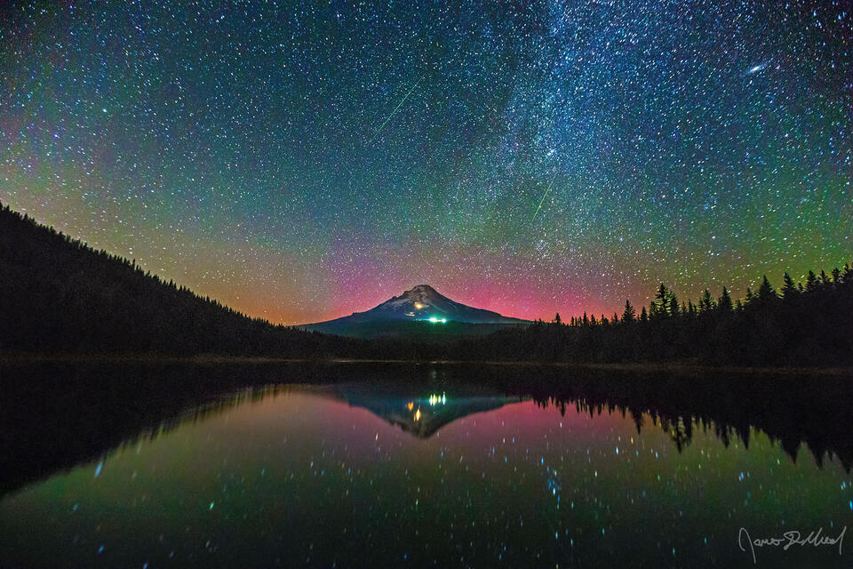 A pink glow from the Aurora Borealis begins to show above Mount Hood. Perseid Meteors shoot across the sky while the scene reflects in Trillium Lake.