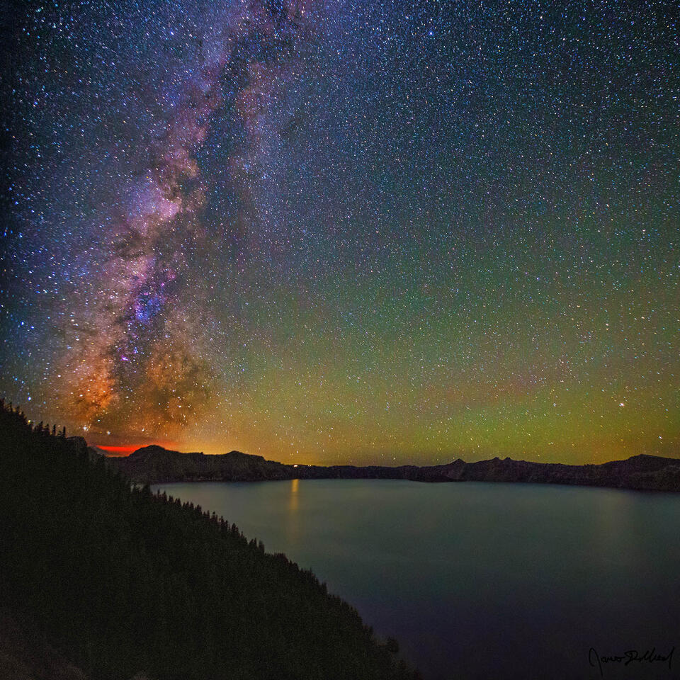 The Milky Way rises high into the night sky above Grotto Cove and Crater Lake at night as s faint airglow illuminates the skies above.