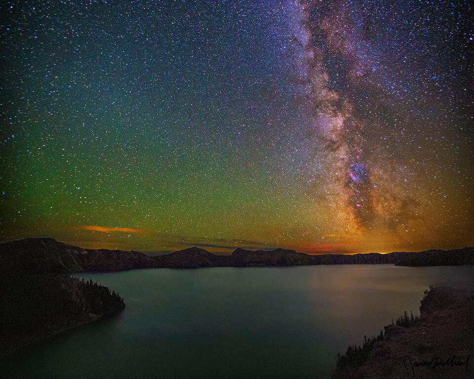 The Milky Way rises high into the night sky above Cleetwood Cove and Crater Lake at night as faint airglow illuminates the skies above.