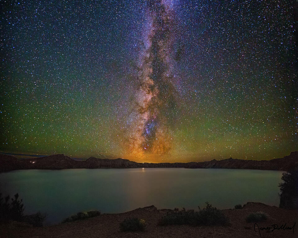 The Milky Way rises high into the night sky above Cleetwood Cove and Crater Lake at night as faint airglow illuminates the skies above.