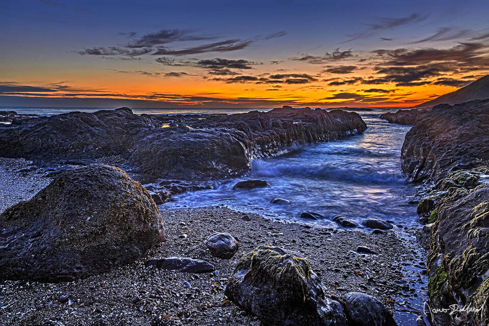 The last colors of sunset hang on over the Pacific Ocean while the rising tide rolls in through a gap in the surrounding rocks as dusk nears.