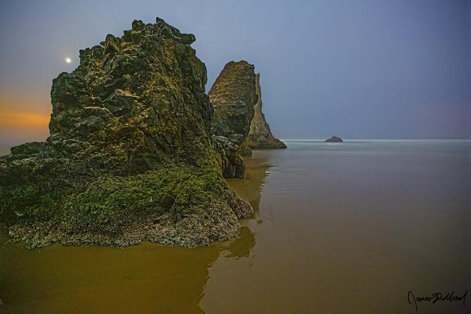 The moon begins to set behind sea stacks in the Pacific Ocean as clouds cover the midnight sky.