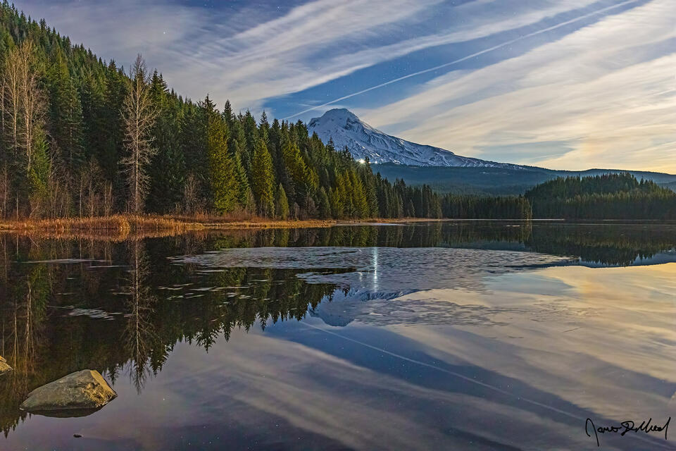 Waning gibbous moonlight illuminating Mount Hood and Trillium Lake at night. Floating ice patches in mirrored water under a streaking cloud sky.