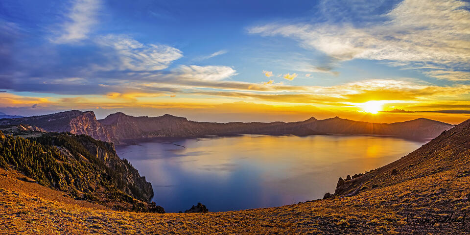 The sun's rays create a warm glow on the Rim of Crater Lake as the colors of sunset reflect off the waters below