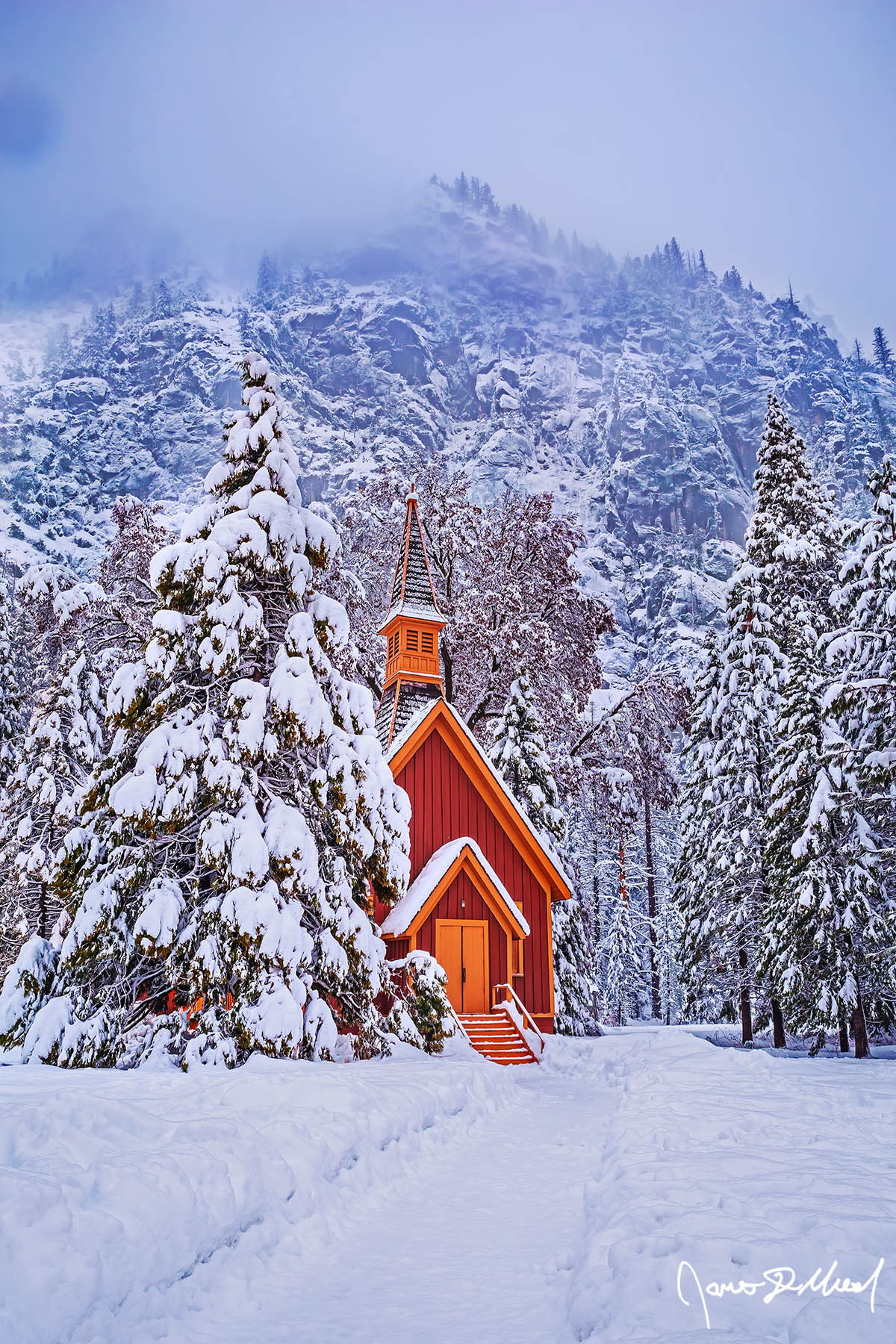 Whisper Snow blankets the Yosemite Valley Chapel, the surrounding trees and meadow at dusk as fog clings to the snow-covered granite cliffs above the Yosemite Valley.