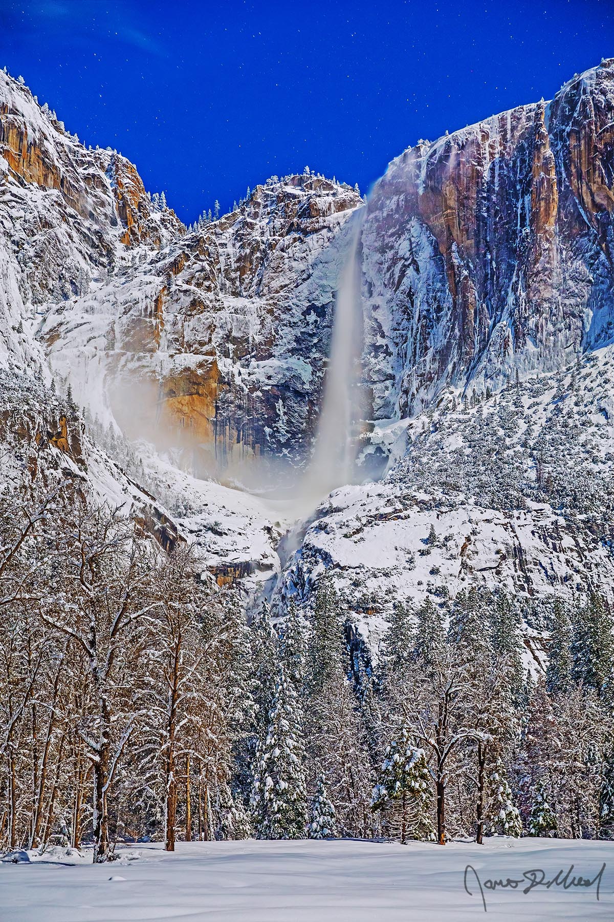 Glimmer Winter moonlight illuminates the night sky, Yosemite Falls and the granite cliffs above Yosemite Valley as snow blankets Cook's Meadow and the valley below.